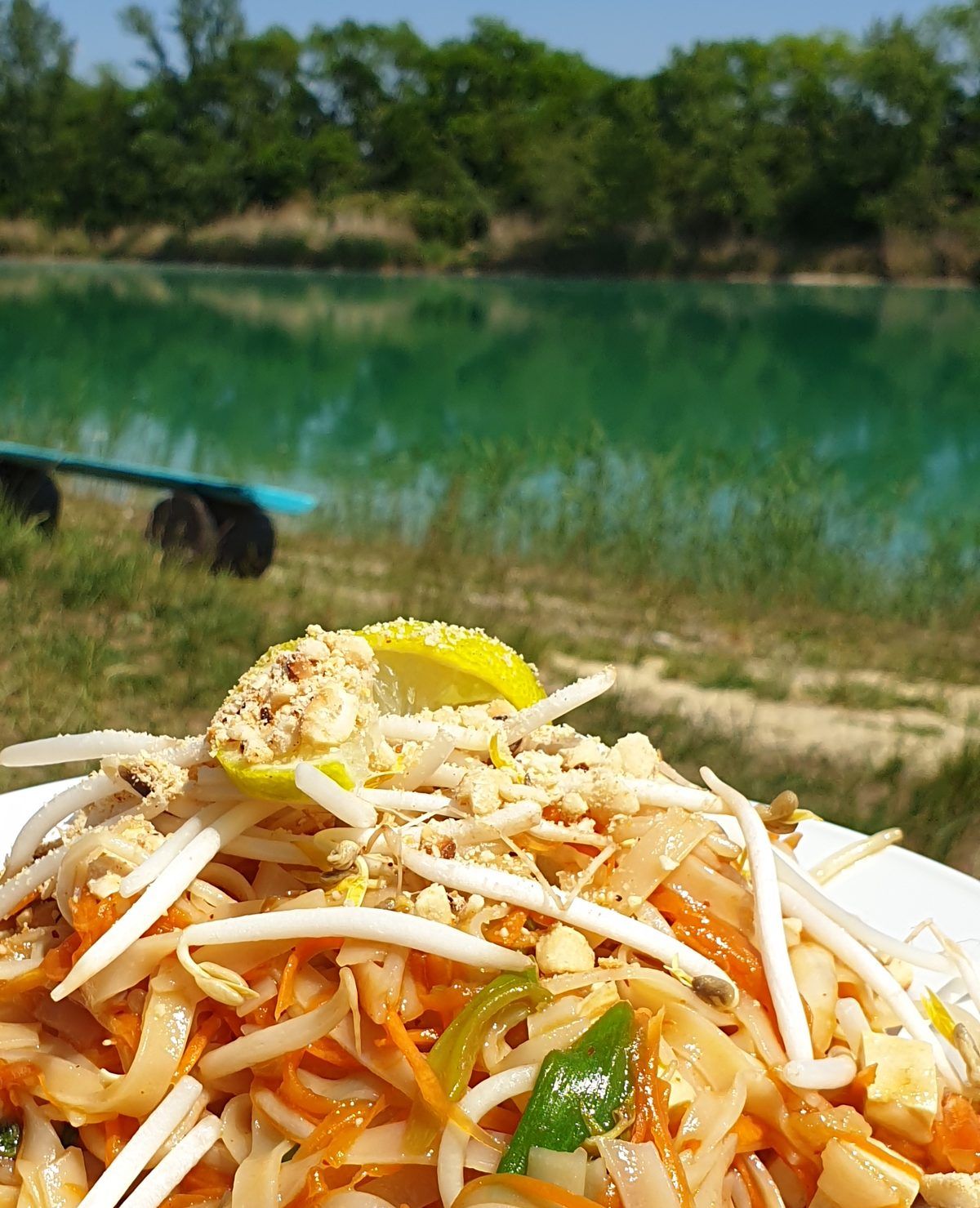 Frisch gekochtes Pad Thai auf einem Teller, dahinter ein türkiser Badesee